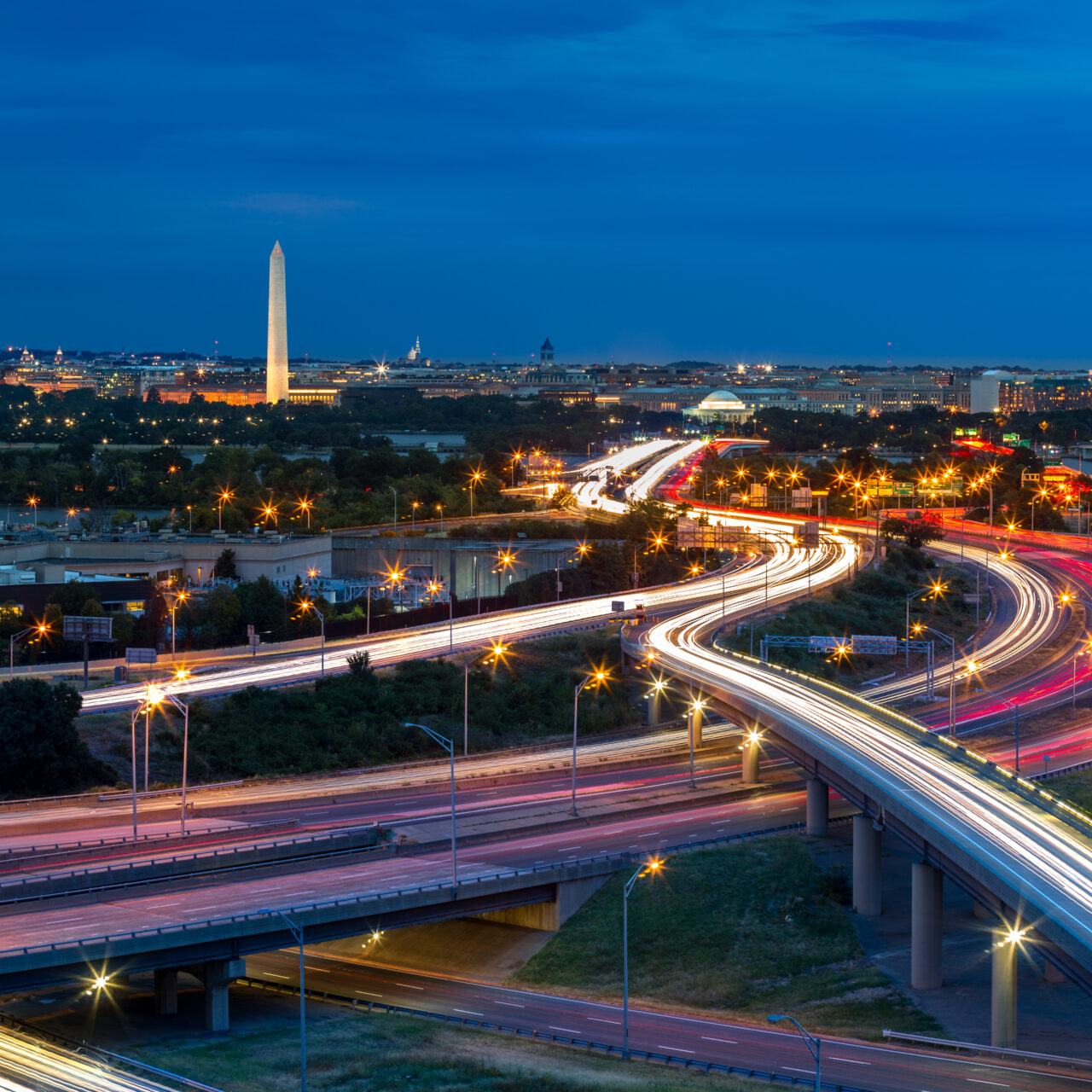 Washington D.C. cityscape at dusk with rush hour traffic trails on I-395 highway. Washington Monument, illuminated, dominates the skyline.